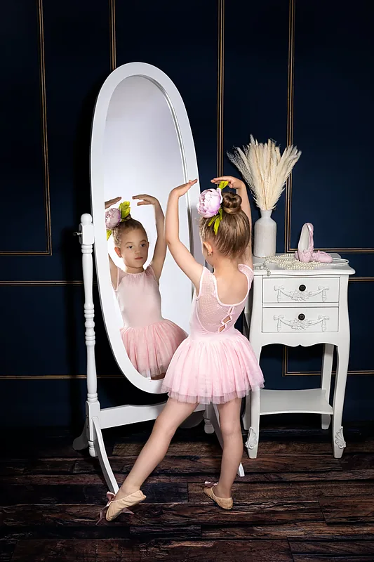 Girl in a tutu looking into a tall oval mirror beside a white dressing table