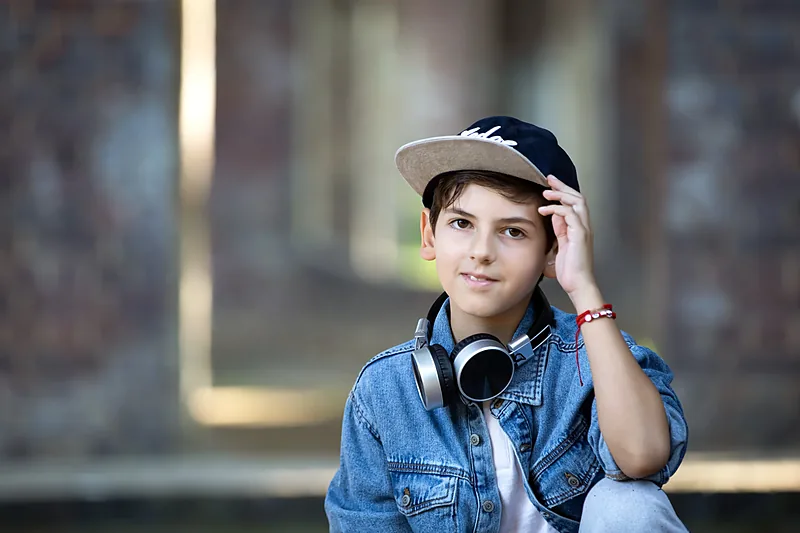 Boy in a denim jacket and cap with headphones around his neck, brick wall behind