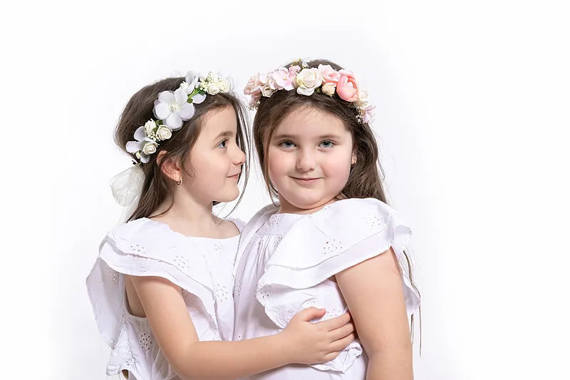 Two girls in white dresses and flower crowns hugging, white background