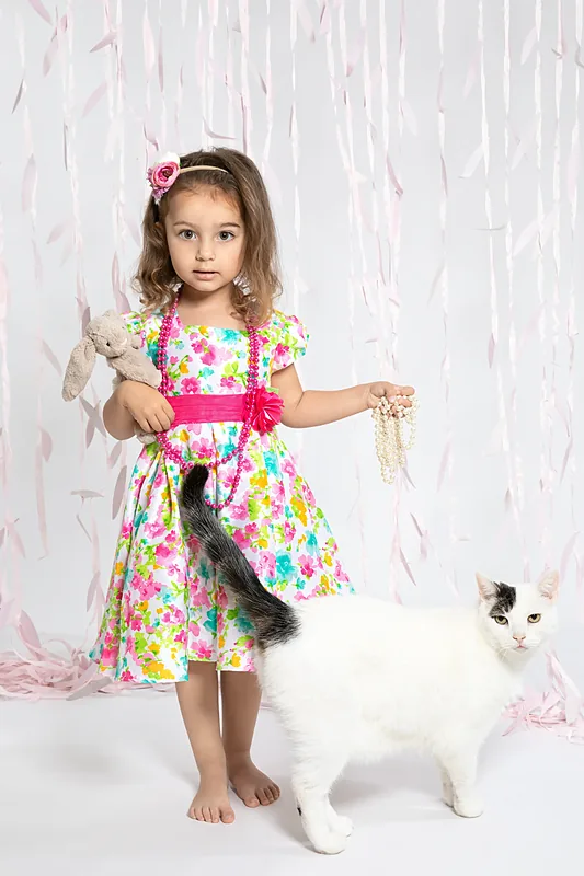 Girl in a bright floral dress standing beside a white cat, white backdrop