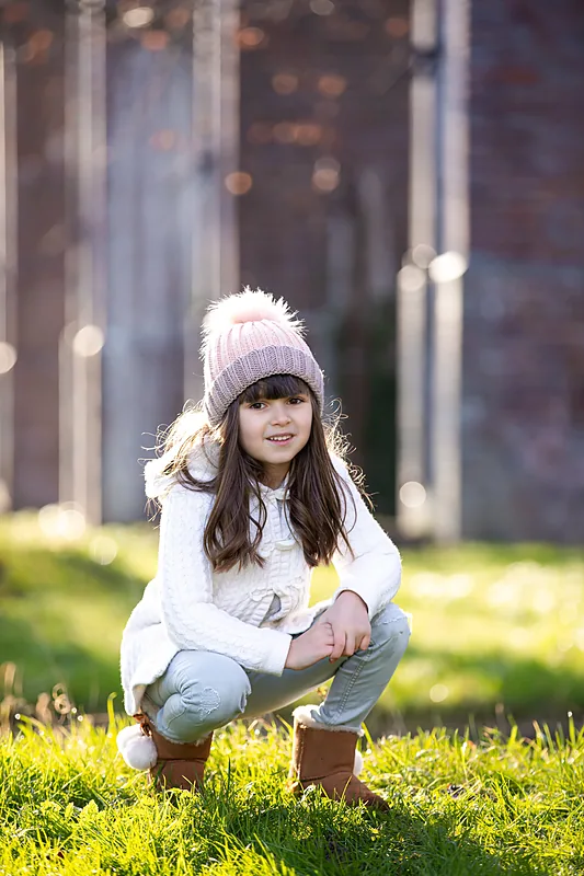 Girl in a pink bobble hat crouching on the grass in front of an old brick building