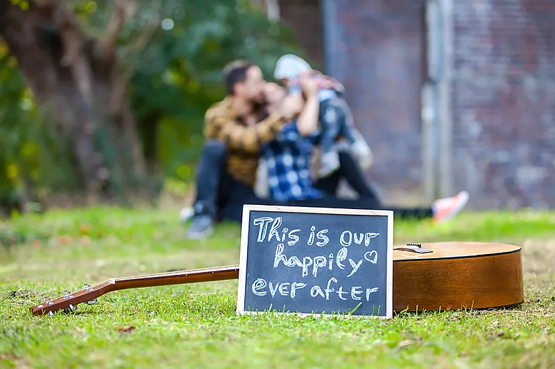 Family with a guitar and a chalkboard sign on the grass beside an old brick building
