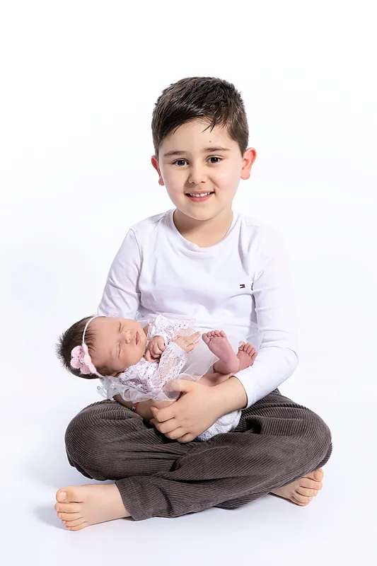 Boy in a white shirt sitting on the floor holding a sleeping newborn