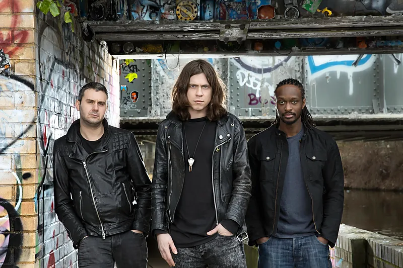 Three musicians in black leather jackets in front of a brick wall