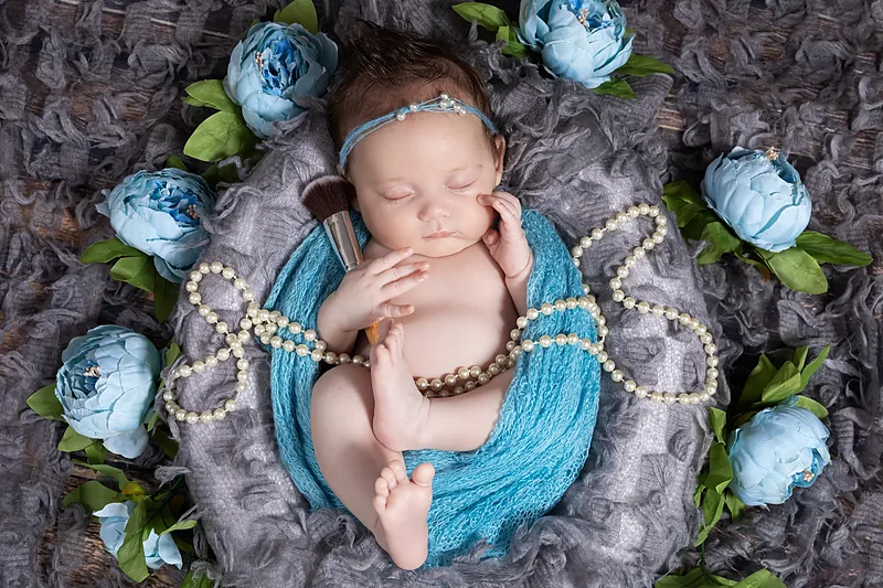 Newborn baby lying on blue and grey fabric with pale blue flowers and a string of pearls