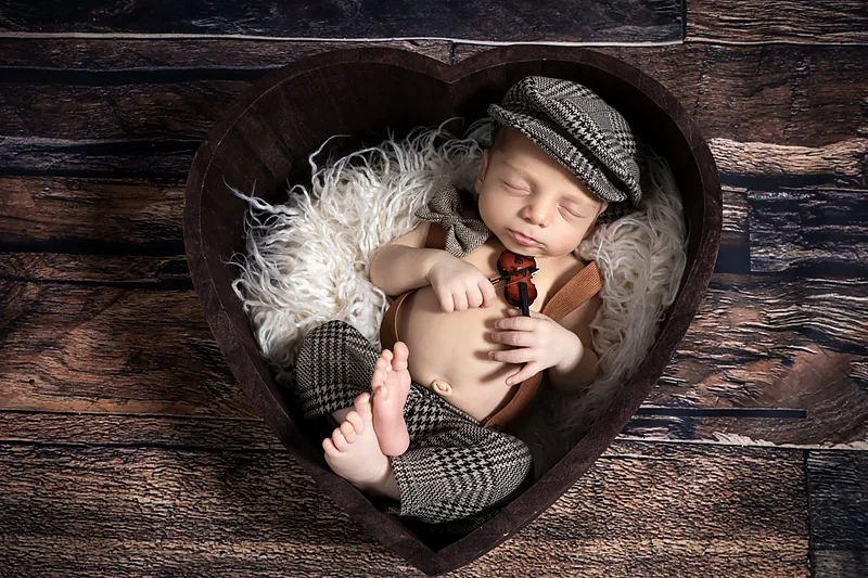 Newborn in a flat cap curled up in a heart-shaped wooden bowl on fur