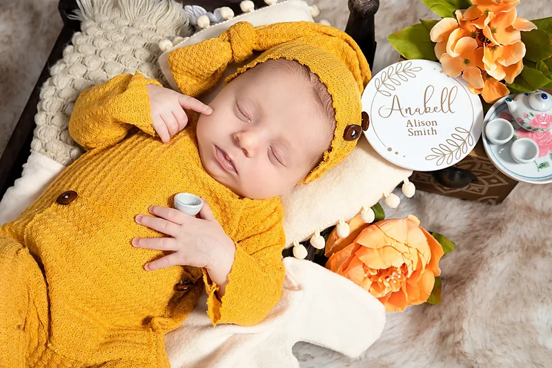 Newborn in a mustard outfit sleeping on white fur beside orange flowers and a small round sign