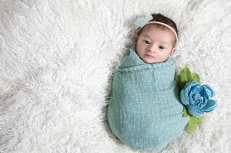Newborn wrapped in a light blue blanket on white fur with a blue flower