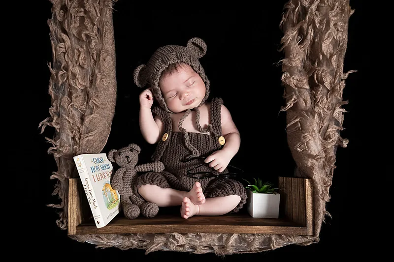 Newborn in a brown bear hat sitting on a wooden swing with a teddy bear