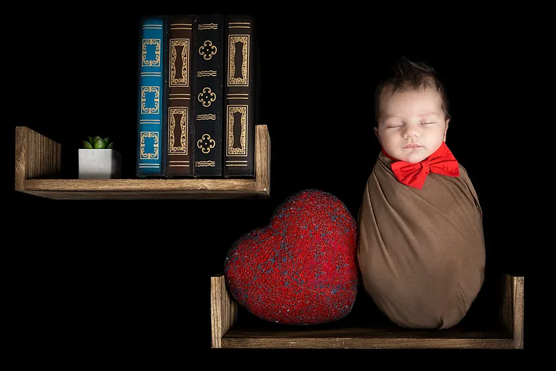 Newborn wrapped in brown with a red bow tie on a wooden shelf with books and a red heart
