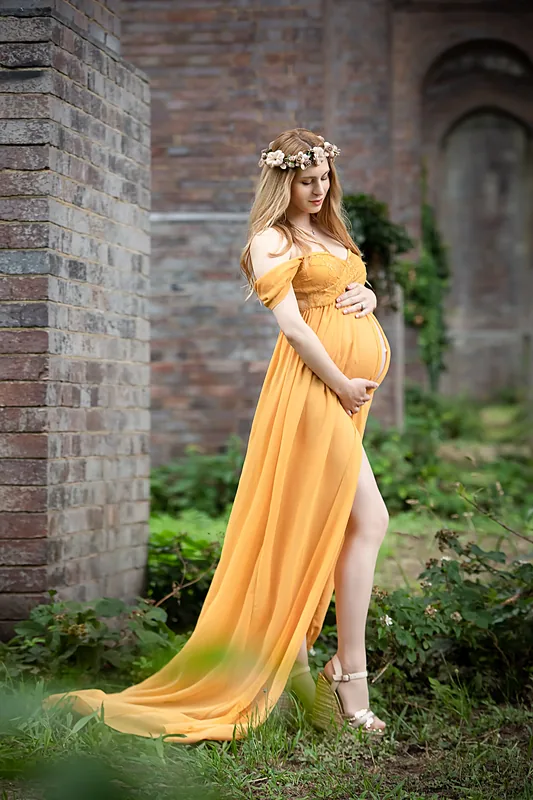 Expectant mother in a flowing yellow gown standing by an ivy-covered brick wall