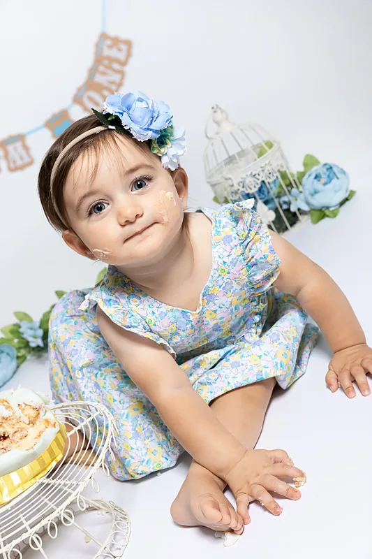 Baby in a blue floral outfit and headband sitting beside a birthday cake