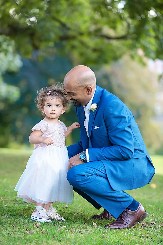 Groom in a blue suit crouching to talk to a small flower girl on the grass