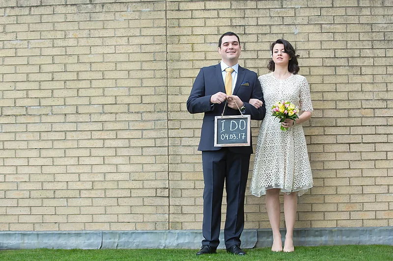 Bride and groom holding a framed sign in front of a pale brick wall