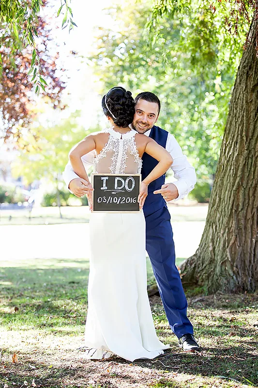 Bride and groom embracing beneath a tree, the groom holding a small sign