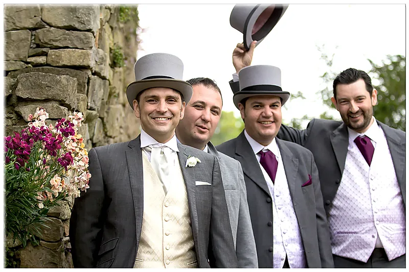 Groom and groomsmen in top hats posing beside a stone wall with flowers